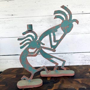 May include: Two metal sculptures featuring stylized figures with textured teal and copper-toned finishes. One figure stands on a base playing a flute, while the other figure stands on a separate base with a small cylindrical candle holder attached.