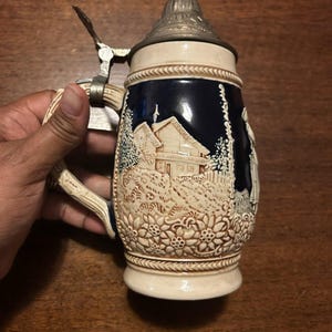 May include: A ceramic beer stein featuring a dark blue body with a raised, cream-coloured relief depicting a house and floral patterns. The stein includes a metallic hinged lid and a curved handle, showcasing detailed craftsmanship and texture.
