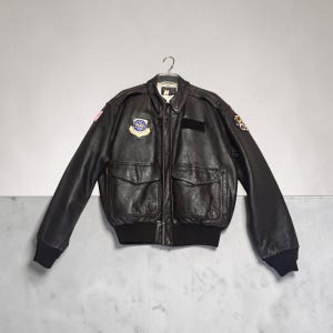 Op de afbeelding: Een donkerbruine leren vliegersjack met een klassieke kraag, zwarte geribbelde boorden bij de mouwen en zoom, en een ritssluiting aan de voorzijde. Het jack is versierd met diverse geborduurde patches op de borst en mouwen, waaronder een rond embleem.