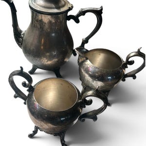 May include: A three-piece silver-plated tea set consisting of a lidded teapot, a creamer, and a sugar bowl. Each piece features a tarnished metallic finish, ornate curved handles, and decorative feet. The teapot has a tall spout and a finial lid.