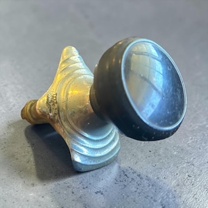 Puede incluir: Un pomo para mueble con una cabeza circular negra unida a una base metálica. La base tiene forma triangular con crestas curvas escalonadas y un acabado en tono dorado desgastado. El pomo está diseñado para montarse en una superficie.