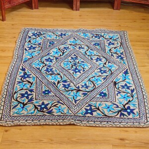 Op de afbeelding: Een vierkant textielkleed met een beige basis en een centraal ruitpatroon. Het ontwerp is versierd met geborduurde bloemmotieven in tinten lichtblauw, donkerblauw en zwart, omlijst door een decoratieve zigzagrand langs de zijkanten.