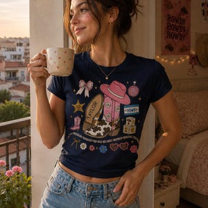 May include: A navy blue short-sleeved t-shirt featuring a graphic print of a pink cowboy hat, a cow-print boot with the name Taylor, a disco ball, a bow, and blocks spelling Howdy. The design includes small flowers, stars, and heart-shaped glasses.