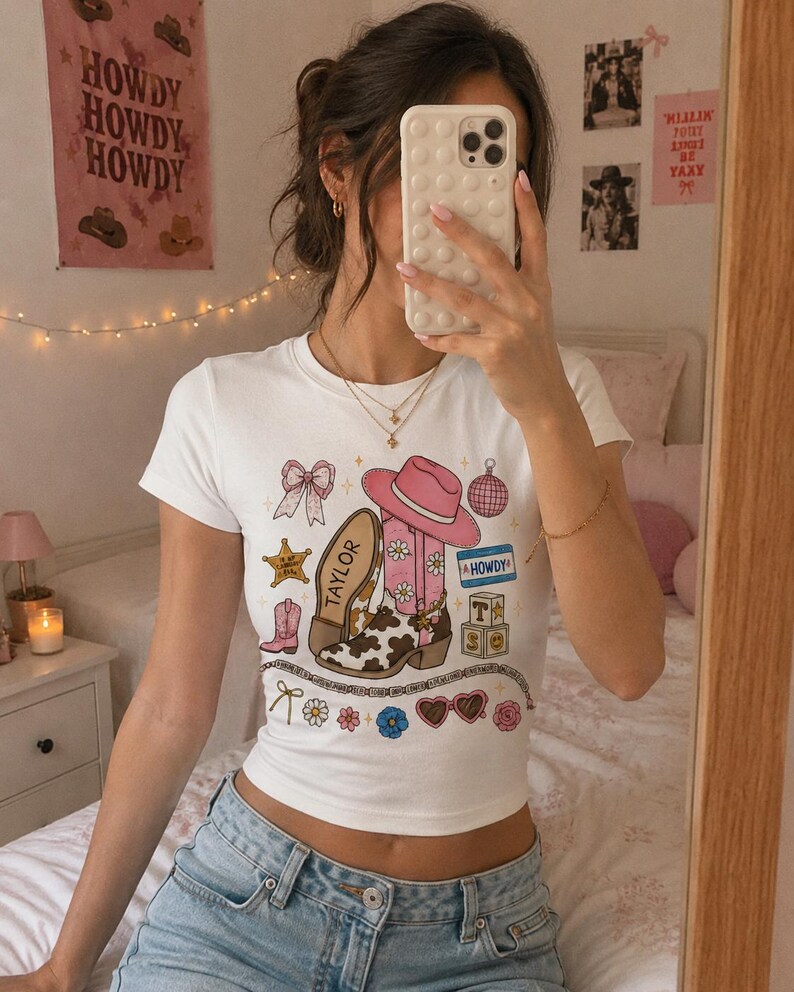 May include: A white short-sleeved t-shirt featuring a graphic print of a pink cowboy hat, a cow-print boot labeled Taylor, a bow, a disco ball, a star, a name tag reading Howdy, alphabet blocks, heart-shaped sunglasses, and various small floral accents.