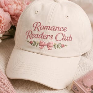 Może przedstawiać: Kremowa czapka z daszkiem z wyhaftowanym napisem Romance Readers Club w kolorze przybrudzonego różu. Poniżej tekstu znajduje się haftowany wzór różowej kokardki otoczonej małymi zielonymi listkami oraz drobnymi różowymi i żółtymi kwiatkami.