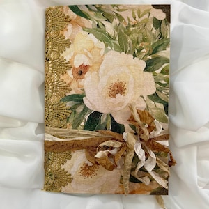 May include: A floral journal with a cream and gold color scheme. The cover features a large peony design with green leaves. The spine is adorned with gold lace trim, and a brown ribbon is tied around the journal.
