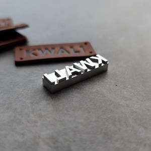 Puede incluir: Un sello de metal plateado con las letras HAWX en relieve, colocado frente a una etiqueta de cuero marrón rectangular con la palabra KWALIT grabada. El sello de metal está diseñado para marcar o estampar materiales.