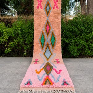 May include: A long, rectangular runner rug featuring a peach-colored upper section and a light pink lower section. The rug displays a central vertical pattern of diamonds in shades of blue, green, orange, and hot pink, finished with white tassels.