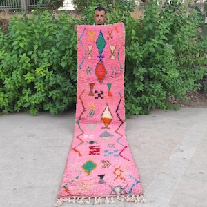 May include: A long, rectangular pink runner rug featuring a variety of colorful geometric shapes and symbols, including diamonds, zigzags, and crosses in green, orange, blue, and black, finished with white fringe tassels at the bottom edge.
