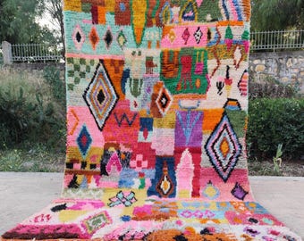 May include: A rectangular area rug featuring a vibrant, multicolored geometric pattern with diamonds, squares, and abstract shapes in shades of pink, orange, yellow, green, blue, and purple. The rug has a thick, textured pile and white fringe.