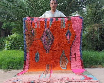 May include: A rectangular hand-woven rug featuring a vibrant orange center with dark blue and pink geometric diamond patterns. The rug has a thick pink border, a light pink base section with multi-colored accents, and white fringe along the bottom edge.