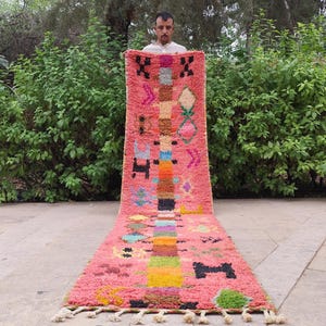 May include: A long, rectangular runner rug featuring a vibrant pink base with a central vertical column of multicolored geometric shapes. The rug is decorated with various black, blue, yellow, and green symbols and has white fringe at the bottom edge.