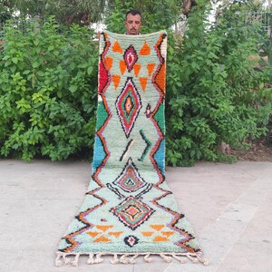 May include: A long, rectangular runner rug featuring a light mint green base with a repeating geometric pattern in orange, red, black, and blue. The design includes diamond shapes and triangular motifs, finished with white fringe at the bottom edge.