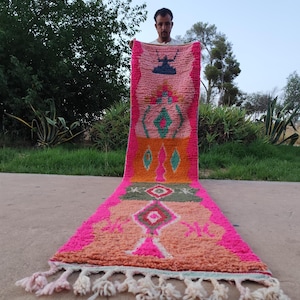 Puede incluir: Una alfombra alargada y rectangular con un patrón geométrico multicolor vibrante en tonos rosa intenso, naranja, melocotón y verde oliva. La alfombra tiene una textura de pelo largo y grueso, rematada con flecos de color crema en los extremos.