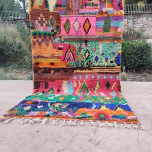 May include: A rectangular hand-knotted wool rug featuring a vibrant, multi-colored geometric pattern. The design includes various shapes like diamonds and triangles in shades of hot pink, orange, green, blue, and brown, finished with white fringe.