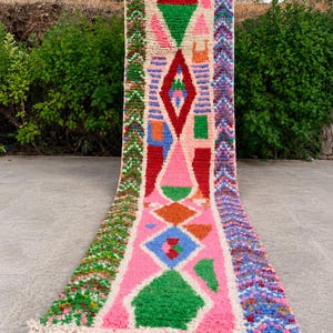 May include: A long, rectangular runner rug featuring a cream-coloured base with a vibrant, multi-coloured geometric pattern. The design includes diamonds, triangles, and textured vertical borders in shades of pink, green, red, blue, and orange, finished with fringe.