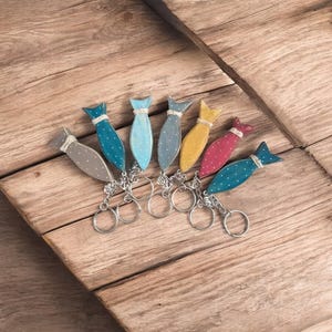 Può includere: Una collezione di sette portachiavi a forma di pesce in legno di vari colori, tra cui grigio, blu, giallo e rosa. Ogni pesce ha un motivo a pois bianchi e un anello portachiavi argentato. I portachiavi sono disposti su una superficie di legno.