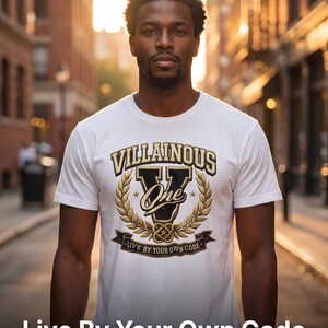 Può includere: Una t-shirt bianca a maniche corte con una grafica centrale che riporta il testo Villainous One, una grande lettera V e un disegno a corona d'alloro. Sotto la corona, un nastro mostra il testo Live By Your Own Code in oro e nero.