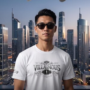 Può includere: Una t-shirt bianca a maniche corte con una stampa grafica sul petto che riporta il testo "VILLAINOUS" sopra un'icona a forma di diamante e il numero "1" all'interno di un bordo rettangolare. La manica mostra il testo "Allteez" in corsivo.