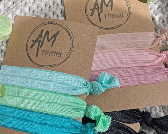 May include: A set of hair scrunchies in a variety of colors, including black, teal, green, and pink. The scrunchies are packaged on a brown card with the logo "AM aideymakes".