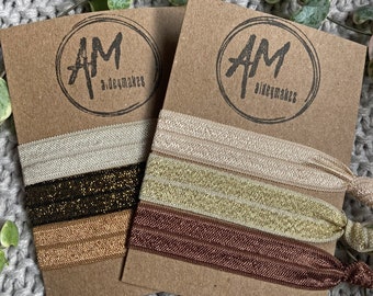 May include: Two sets of hair scrunchies on brown card stock. Each set has three scrunchies in different colors and textures. The scrunchies are made of elastic fabric with a shimmery finish. The card stock has the logo "AM a.deuxmakes" printed on it.