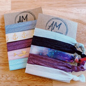 May include: Two sets of hair scrunchies on brown cardstock. The scrunchies are made of fabric and have different patterns. One set has a mermaid, anchors, and a galaxy print. The other set has a galaxy print, a mermaid, and a solid color. The cardstock has the text "AM" on it.