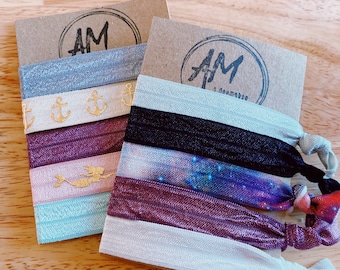 May include: Two sets of hair scrunchies on brown cardstock. The scrunchies are made of fabric and have different patterns. One set has a mermaid, anchors, and a galaxy print. The other set has a galaxy print, a mermaid, and a solid color. The cardstock has the text "AM" on it.