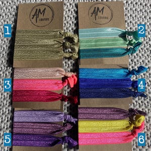 May include: Six sets of colorful hair ties with glitter. Each set contains three hair ties in different shades of the same color. The hair ties are arranged in rows on a brown background with the text "AM Accessories" printed on each card.