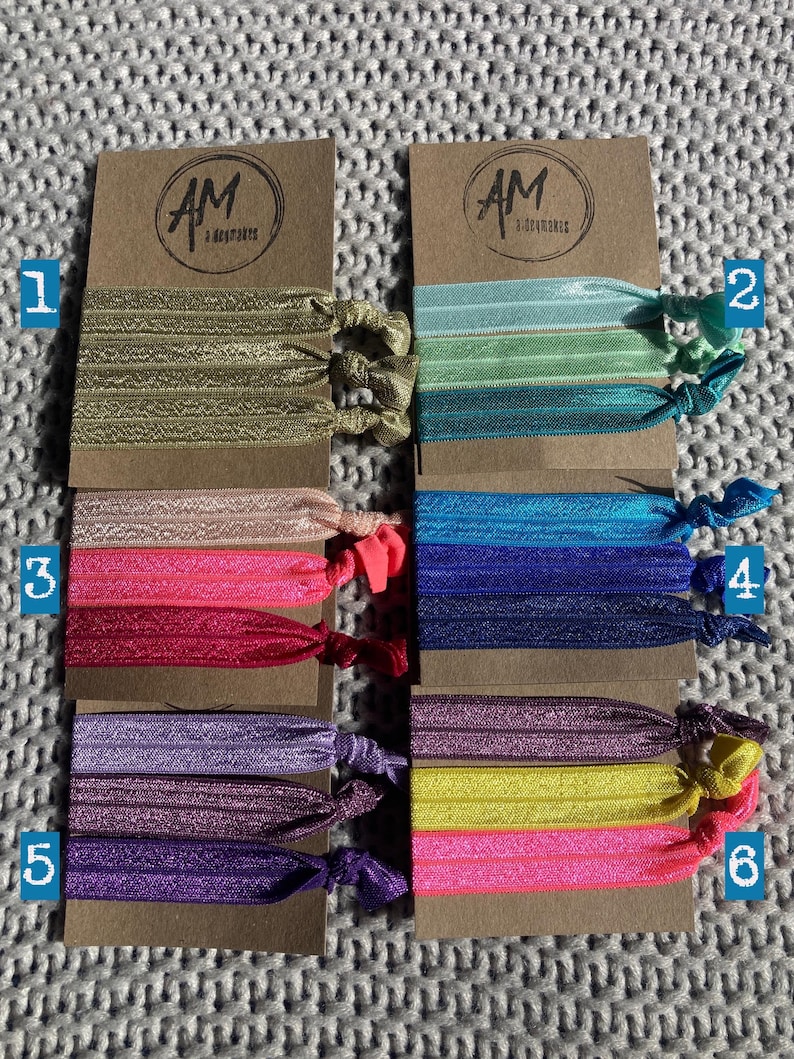 May include: Six sets of colorful hair ties with glitter. Each set contains three hair ties in different shades of the same color. The hair ties are arranged in rows on a brown background with the text "AM Accessories" printed on each card.