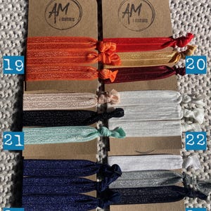 May include: Set of 10 colorful hair ties, packaged in pairs on brown cards with the text "AM Accessories". The hair ties are made of elastic and come in a variety of colors, including orange, red, gold, burgundy, beige, black, light blue, white, silver, and navy blue.