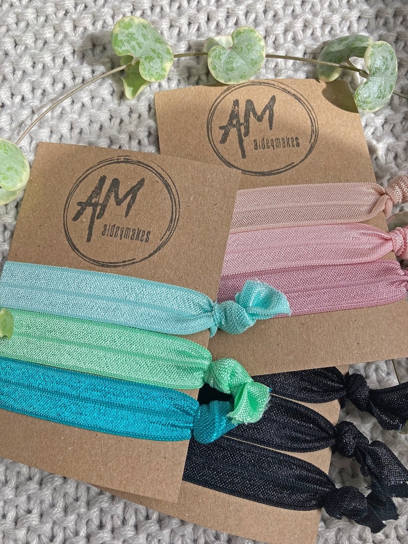 May include: A set of hair scrunchies in a variety of colors, including black, teal, green, and pink. The scrunchies are packaged on a brown card with the logo "AM aideymakes".