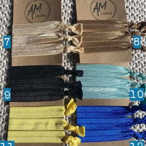 May include: Set of 6 hair scrunchies in various colors, including black, beige, yellow, blue, and light blue. Each scrunchie is made of a soft, stretchy fabric and comes in a set of three. The scrunchies are displayed on a brown card with the text "AM Accessories" printed on it.