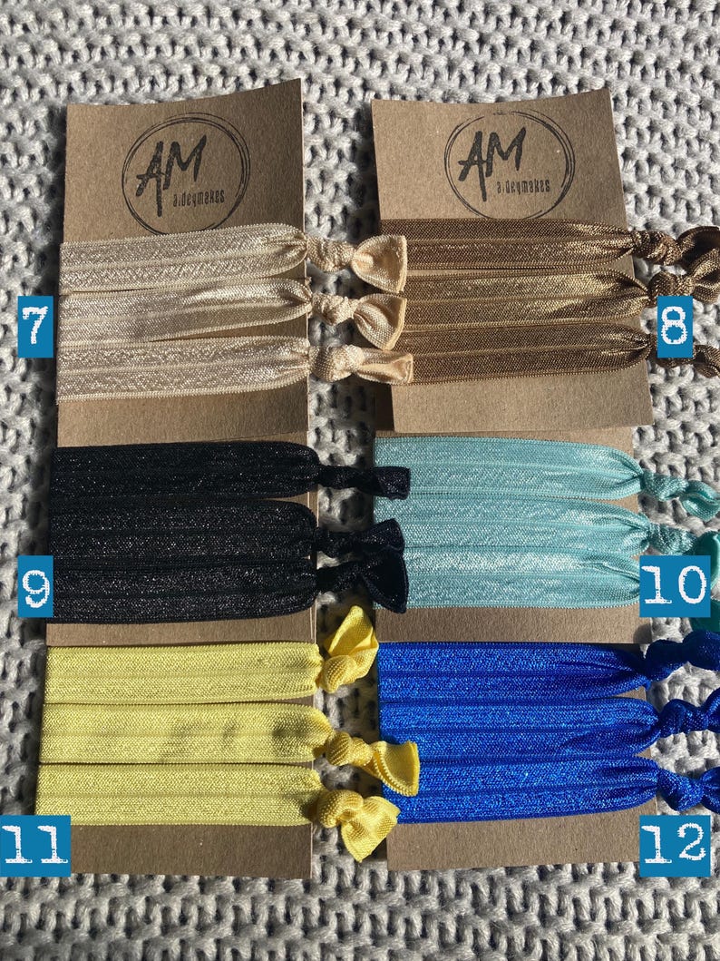 May include: Set of 6 hair scrunchies in various colors, including black, beige, yellow, blue, and light blue. Each scrunchie is made of a soft, stretchy fabric and comes in a set of three. The scrunchies are displayed on a brown card with the text "AM Accessories" printed on it.