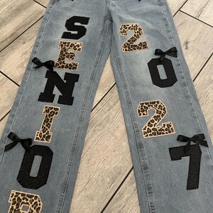 May include: Light blue denim jeans featuring the word SENIOR on the left leg and 2027 on the right leg. The letters and numbers alternate between a black glitter finish and a leopard print pattern. Small black satin bows are attached throughout.