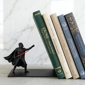 Op de afbeelding: Een zwarte metalen boekensteun met een gebeeldhouwd Darth Vader-figuur die een rood lichtzwaard vasthoudt, ondersteunt vijf boeken met de titels Dune, Star Wars: Archivi Completi, I Segreti della Galassia, Teoria della Forza en Enciclopedia della Saga di Skywalker.