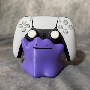 Könnte beinhalten: Ein violetter Ditto-förmiger Controller-Ständer, der einen weißen und schwarzen PlayStation 5-Controller hält. Der Ständer hat ein einfaches, lächelndes Gesicht. Der Controller hat das PlayStation-Logo und die Tasten-Symbole.