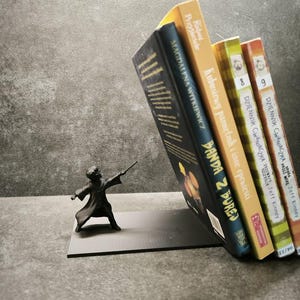 Könnte beinhalten: Eine schwarze Zaubererfigur mit Zauberstab, die einen Stapel bunter polnischer Bücher hält. Der Zauberer steht auf einer grauen Basis. Der Hintergrund ist eine strukturierte graue Oberfläche. Die Bücher sind auf Polnisch.