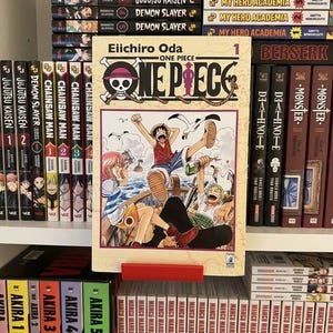Peut inclure: Un volume 1 du manga One Piece par Eiichiro Oda avec une illustration de couverture montrant trois personnages sur le pont d'un navire et un logo tête de mort. Le livre est présenté sur une étagère entouré d'autres volumes de mangas.