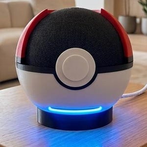 Könnte beinhalten: Ein kugelförmiger Smart-Lautsprecher, der wie ein rot-weiß-schwarzer Ball gestaltet ist. Das Gerät verfügt über einen oberen Bereich aus schwarzem Stoffgewebe, eine weiße untere Hälfte, einen zentralen runden Knopf und einen leuchtenden blauen Lichtstreifen am Sockel.