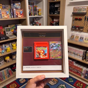 May include: A white shadow box frame containing a red Pokemon Red Version Game Boy game cartridge and a Charizard trading card. The display features a red patterned background with text labels including Pokemon Red Version and a Charizard stat box.