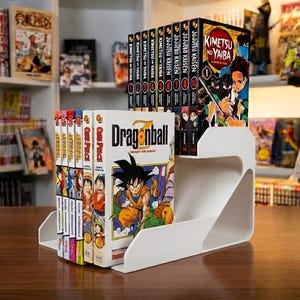 Puede incluir: Un expositor blanco de dos niveles que contiene una colección de libros manga. El nivel inferior muestra volúmenes de Dragonball y One Piece, mientras que el nivel superior exhibe una serie de mangas de Jujutsu Kaisen y Kimetsu no Yaiba con portadas ilustradas.