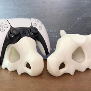 Op de afbeelding: Een wit en zwart draadloze gamecontroller rustend op een crèmekleurige schedelvormige standaard, naast een identieke lege standaard. De controller is voorzien van een centraal touchpad, twee analoge sticks en diverse richtingsknoppen.