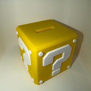 Può includere: Un salvadanaio cubico giallo con un simbolo di punto interrogativo bianco sulle facce anteriore e laterale. La superficie superiore include una fessura rettangolare per le monete e piccoli accenti circolari bianchi agli angoli.