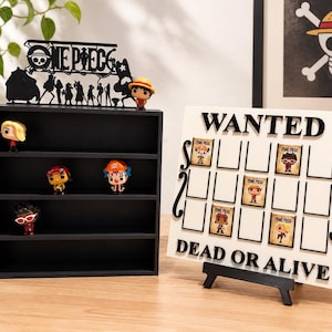 Può includere: Uno scaffale espositivo nero a quattro ripiani con un logo One Piece sulla parte superiore e una tavola rettangolare bianca con il testo WANTED DEAD OR ALIVE con spazi vuoti per piccole carte personaggio, insieme a varie statuine.