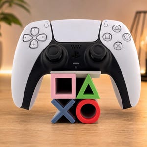 Peut inclure: Une manette de jeu sans fil blanche et noire posée sur un support en forme des quatre symboles PlayStation : un carré rose, un triangle vert, une croix bleue et un cercle rouge. La manette dispose d'un pavé tactile central et de deux joysticks.