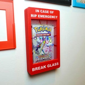 Könnte beinhalten: Ein rechteckiger, roter Wand-Display-Kasten mit einer Pokemon Prismatic Evolutions Sammelkartenspiel-Packung. Der Rahmen ist mit dem weißen Text IN CASE OF RIP EMERGENCY und BREAK GLASS versehen, der an einen Notfallkasten erinnert.
