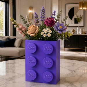 Pode incluir: Um vaso retangular roxo em forma de um grande bloco de LEGO com seis pinos circulares na frente, cada um gravado com o logotipo da LEGO. O vaso está preenchido com um buquê colorido de flores artificiais, incluindo lavanda, rosas e flores roxas.