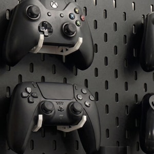 Puede incluir: Un panel perforado negro con soportes de montaje de plástico blanco que sostienen varios mandos de videojuegos negros. Los mandos incluyen un modelo Xbox con botones de colores y un modelo PlayStation con botones de color negro.