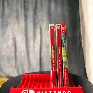 Può includere: Tre custodie di giochi per Nintendo Switch posizionate verticalmente in un supporto in plastica rossa. Le custodie sono Animal Crossing: New Horizons, Pokémon: Let's Go, Eevee! e Pokémon Pokopia. Il supporto presenta il logo Nintendo Switch bianco.