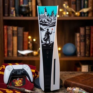 Puede incluir: Una consola de juegos blanca con un gráfico en blanco y negro de un castillo y una figura sosteniendo una varita roja brillante sobre un fondo verde azulado. La consola incluye un mando inalámbrico, una varita y una botella de poción.